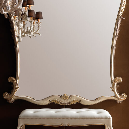 A Large Rococo Style Wall Mirror with an intricately carved frame is mounted on a brown wall. A white cushioned bench sits below it. To the left, a decorative wall sconce with black lampshades adds elegance to the scene.