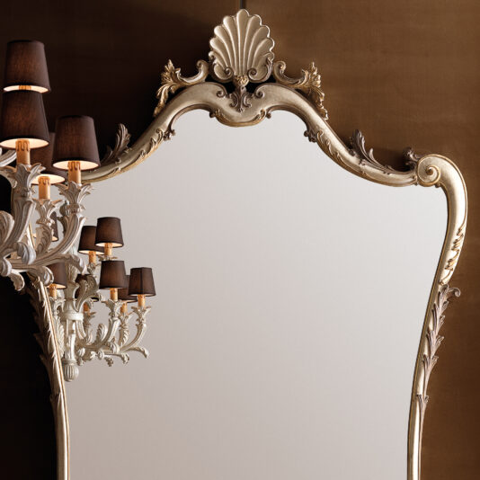 A Large Rococo Style Wall Mirror with an elaborate gold frame and detailed craftsmanship is mounted on a brown wall. A decorative wall sconce featuring five dark lampshades is positioned on the left side of the mirror, casting warm light onto its surface.
