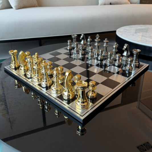 The Luxury Chess Set is elegantly displayed on a reflective glass table, its gold and silver pieces ready for play. In the background, a modern white sofa enhances the scene alongside a round marble-topped side table.