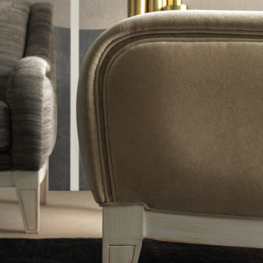 Close-up of two stylish Modern Deco Armchairs with luxurious upholstery. The chair in the foreground showcases a smooth taupe fabric, whereas the one in the background displays a textured gray finish. Both are supported by light wood legs and are positioned against an elegant, minimalist backdrop.