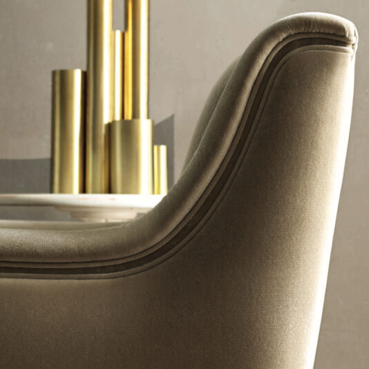 A close-up of the plush, beige Modern Deco Armchair with its gracefully curved edges, situated beside a sleek table. On the table rest several tall, cylindrical gold vases that provide a striking contrast to the armchair's subtle, muted tones. The softly lit background enhances the textures beautifully.