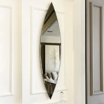 The Modern Pointed Oval Bronze Wall Mirror beautifully captures a portion of the living room, highlighting a sofa and abstract artwork. With its sharp top and bottom, this elongated mirror is installed on a white paneled wall, enhancing the space with contemporary flair.