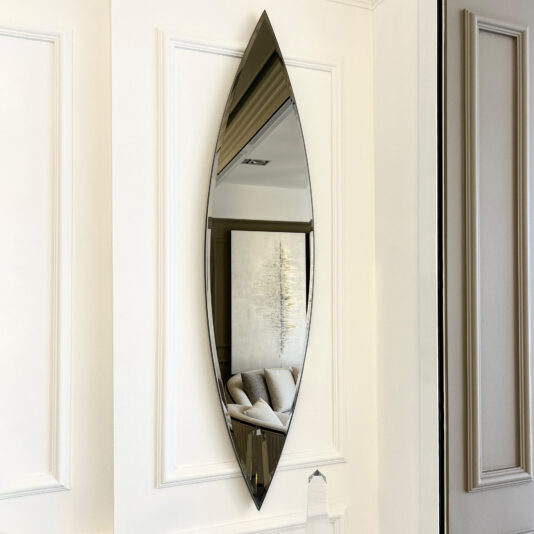 The Modern Pointed Oval Bronze Wall Mirror beautifully captures a portion of the living room, highlighting a sofa and abstract artwork. With its sharp top and bottom, this elongated mirror is installed on a white paneled wall, enhancing the space with contemporary flair.