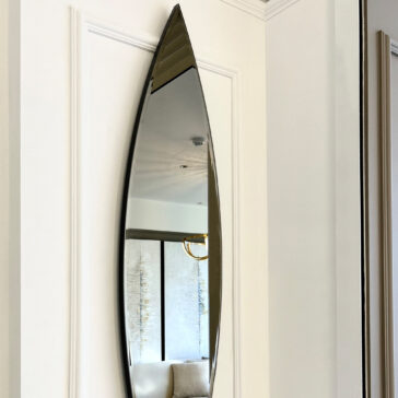 A Modern Pointed Oval Bronze Wall Mirror is mounted on a white wall, showcasing its surfboard-shaped design as it reflects part of a room adorned with abstract artwork and a gold chandelier.