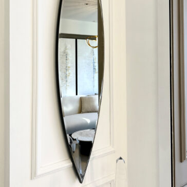 The Modern Pointed Oval Bronze Wall Mirror is elegantly displayed on a pristine white wall, capturing the reflection of a stylish living room featuring a beige couch and a circular marble coffee table. In the mirrored view, large windows softly illuminate the room through sheer curtains.