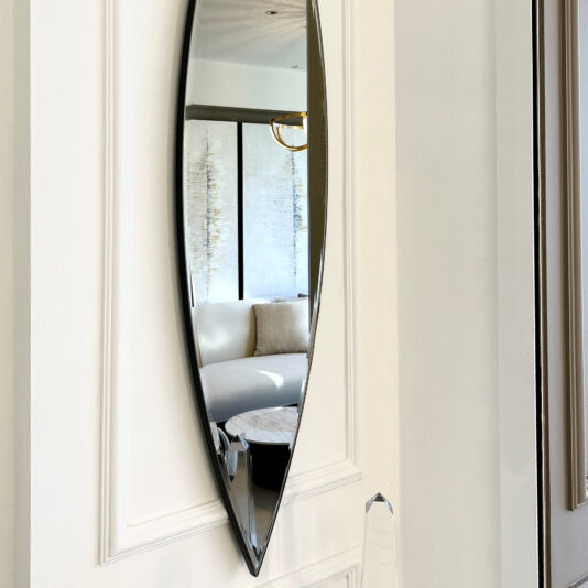 The Modern Pointed Oval Bronze Wall Mirror is elegantly displayed on a pristine white wall, capturing the reflection of a stylish living room featuring a beige couch and a circular marble coffee table. In the mirrored view, large windows softly illuminate the room through sheer curtains.