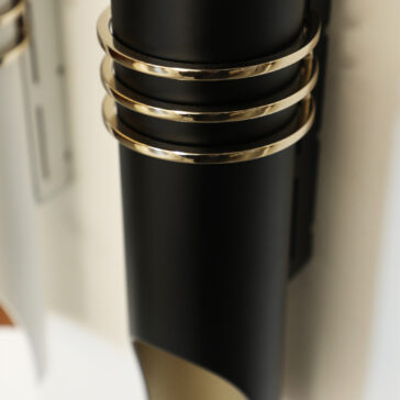 A close-up shot of the Modern Vertical Wall Light showcases a sleek black cylindrical dispenser accented with three silver metal rings, mounted on a wall in a contemporary and minimalistic design.