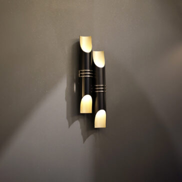The Modern Vertical Wall Light consists of two cylindrical fixtures in black and gold, mounted on a dark gray wall, casting a warm glow from both ends.