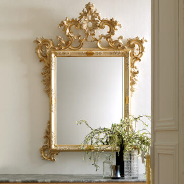 A Rococo Style Rectangular Wall Mirror with an elaborate gold frame and intricate designs is mounted on a white wall. Beneath it, a small table decorated with decorative glass bottles and a flowering plant completes the elegant setting. To the right, part of a door frame is visible.