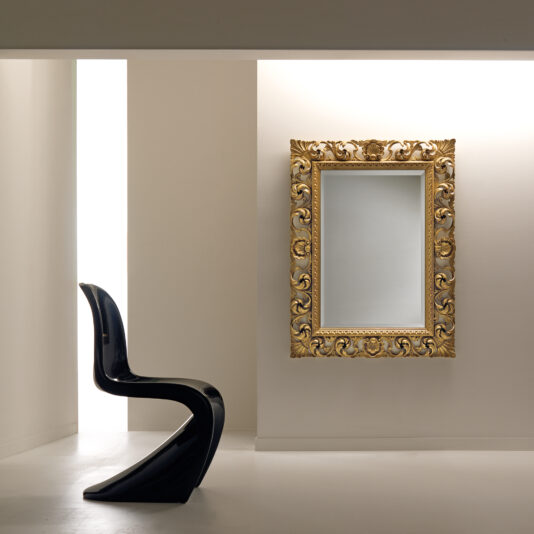 A modern, curved black chair is positioned in front of a wall graced by an Ornate Baroque Style Wall Mirror with an intricately designed gold frame. Soft, natural light filters through a tall, narrow window on the left, highlighting the minimalist setting.