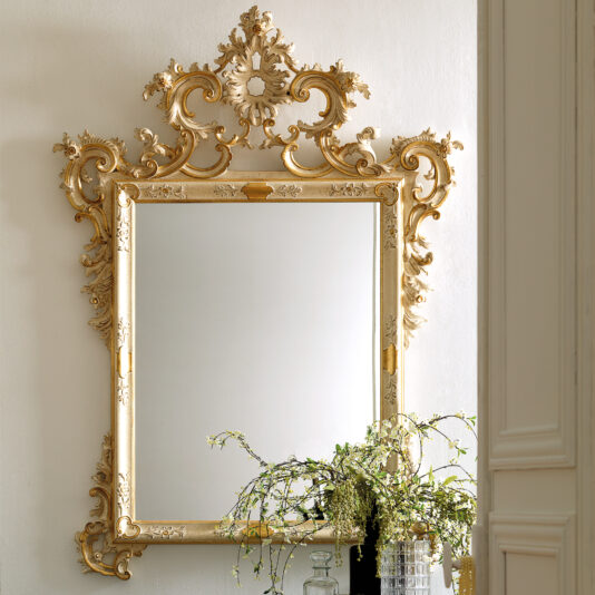 An Baroque Style Wall Mirror with elaborate floral carvings hangs on a wall. Below it, a table holds a vase with delicate, leafy branches.
