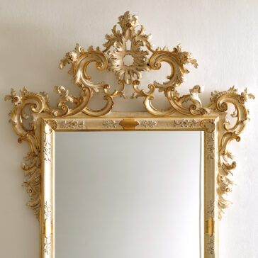 An Ornate Baroque Style Wall Mirror featuring an intricately designed gold frame with floral and scroll motifs, complete with an elaborate crest, adorns a plain white wall. The reflection in the mirror is not visible.