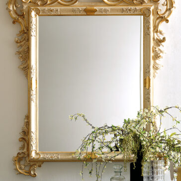 An Ornate Baroque Style Wall Mirror with a gold frame is elegantly displayed on the wall. Beneath it, a glass vase contains a lush arrangement of cascading green foliage, enhancing the decor with an elegant touch.