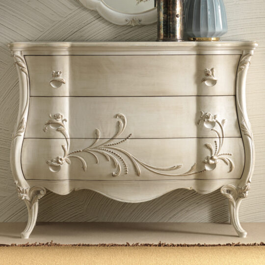 A vintage-inspired Ornate Bombe Chest Of Drawers in a cream hue, adorned with intricate floral carvings on its front and featuring three roomy drawers alongside graceful curved legs, set against a subtly textured wall.