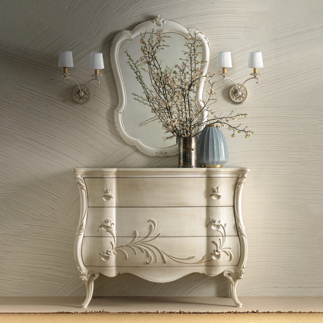 The Ornate Bombe Chest Of Drawers, adorned with vintage-style floral carvings, is elegantly topped with a vase of branches. Above it hangs a large decorative mirror, complemented by two elegant wall sconces that provide soft lighting. The textured wall design further enhances the charming ambiance.