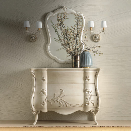 Ornate Bombe Chest Of Drawers The Ornate Bombe Chest Of Drawers, adorned with vintage-style floral carvings, is elegantly topped with a vase of branches. Above it hangs a large decorative mirror, complemented by two elegant wall sconces that provide soft lighting. The textured wall design further enhances the charming ambiance.