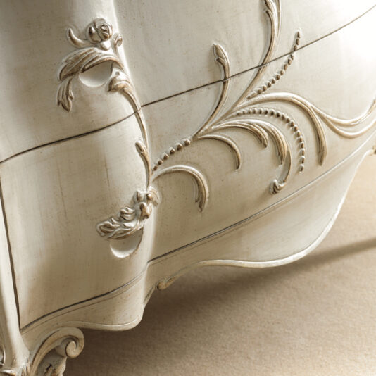 Ornate Bombe Chest Of Drawers A close-up view of the Ornate Bombe Chest Of Drawers shows its vintage white design, featuring intricate floral carvings on its elegantly curved surface. The soft lighting beautifully highlights the texture and detailed woodwork.
