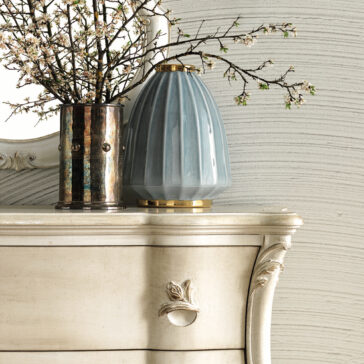 An Ornate Bombe Chest Of Drawers in a vintage-style cream finish, adorned with intricate carvings, supports a ribbed blue vase and a metal container brimming with blooming branches. The backdrop showcases a textured, wavy wall alongside a partial view of a round mirror.