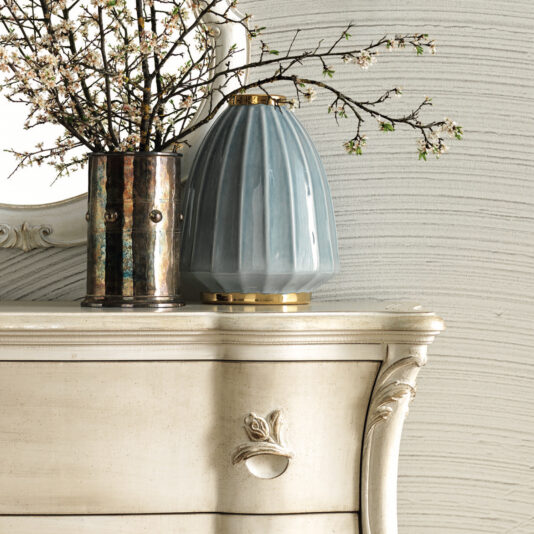 Ornate Bombe Chest Of Drawers An Ornate Bombe Chest Of Drawers in a vintage-style cream finish, adorned with intricate carvings, supports a ribbed blue vase and a metal container brimming with blooming branches. The backdrop showcases a textured, wavy wall alongside a partial view of a round mirror.