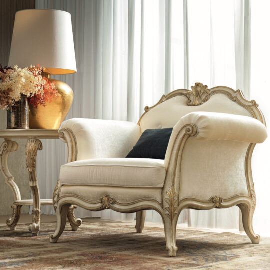 Placed beside a decorative side table featuring a gold lamp and a bouquet of dried flowers, the Ornate Luxury Rococo Style Sofa exudes the elegance of Rococo design with its vintage charm, featuring intricate wooden carvings and cream upholstery set against sheer white curtains.