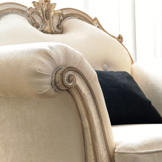 A close-up of an elegant Ornate Luxury Rococo Style Sofa showcases its vintage beige design with exquisite detailing and rolled armrests. A dark pillow adorns the plush backrest, exuding luxury as soft natural light filters through curtains in the background.