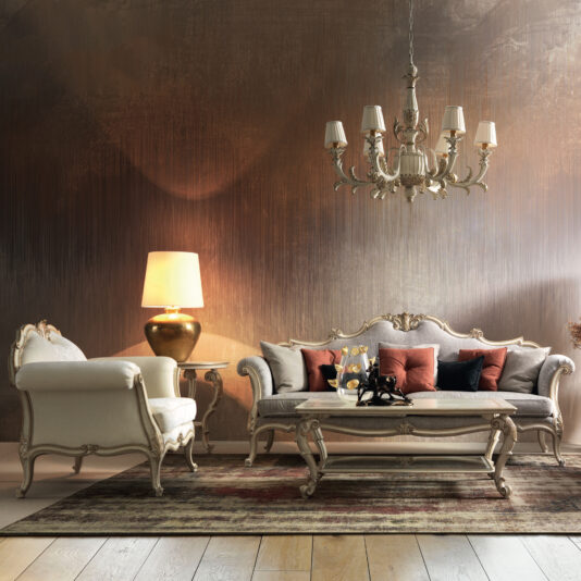 An Ornate Luxury Rococo Style Sofa enhances the opulent living room, paired with an elegant armchair and coffee table. Overhead, a chandelier provides illumination while a warm lamp sits nearby. The richly textured walls and patterned rug on the wooden floor beautifully complement the neutral and dark red tones of this lavish space.