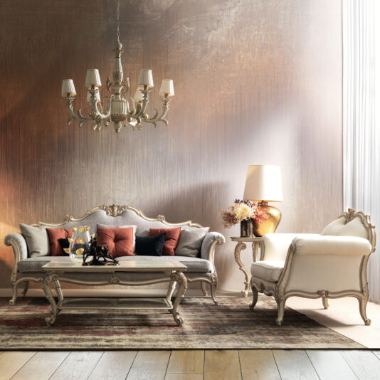A sophisticated living room with a Rococo-style chandelier hangs above an Ornate Luxury Rococo Style Sofa in gray, accompanied by a white armchair and coffee table. The space is adorned with autumn-colored cushions, a table lamp, and a vase of flowers, set against a textured wall and natural wood flooring.