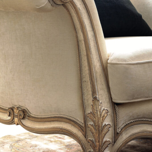 A close-up of the Ornate Luxury Rococo Style Sofa reveals the elegance of its beige upholstery, featuring intricately carved wooden details and a plush cushioned seat. The armrest is adorned with delicate leaf patterns, while a dark pillow peeks out partially from behind. This exquisite piece is complemented by a light-patterned rug beneath it.
