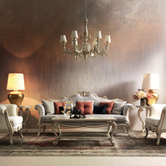 Ornate Luxury Rococo Style Sofa A classic living room featuring the Ornate Luxury Rococo Style Sofa in beige, adorned with red and gray cushions, flanked by two armchairs. A vintage chandelier hangs overhead, complemented by warm table lamps on each side. The setting is completed with a textured wall and a patterned rug, exuding cozy luxury.