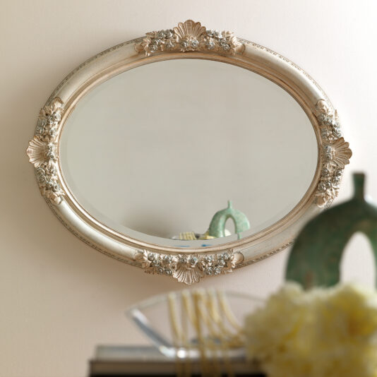 The Oval Rococo Style Mirror, featuring an elaborate frame, is mounted on a light-colored wall. Its reflection captures a portion of a decorative green vase and the soft focus of a yellow flower arrangement in the foreground.