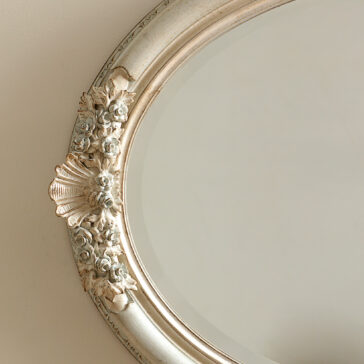 Oval Rococo Style Mirror An up-close view of the Oval Rococo Style Mirror, adorned with an antique finish that highlights its intricate floral and shell designs, is mounted on a plain beige wall.
