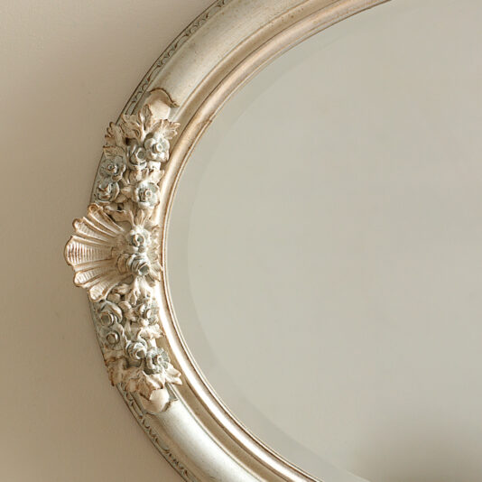 Oval Rococo Style Mirror An up-close view of the Oval Rococo Style Mirror, adorned with an antique finish that highlights its intricate floral and shell designs, is mounted on a plain beige wall.