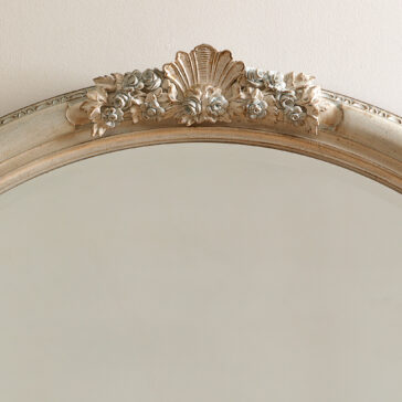 Oval Rococo Style Mirror The Oval Rococo Style Mirror features a faded gold frame embellished with ornamental flowers and a shell at the top, reflecting a soft beige wall.