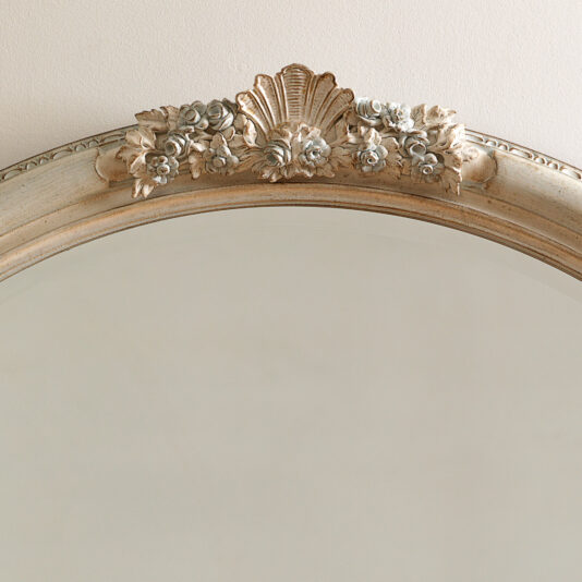 Oval Rococo Style Mirror The Oval Rococo Style Mirror features a faded gold frame embellished with ornamental flowers and a shell at the top, reflecting a soft beige wall.