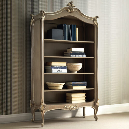 The Rococo Inspired Bookcase, adorned with intricate carvings, showcases a collection of neatly arranged books and decorative bowls. This vintage-style piece features a curved top and elegant details, elegantly standing against a textured wall on a light-colored floor.