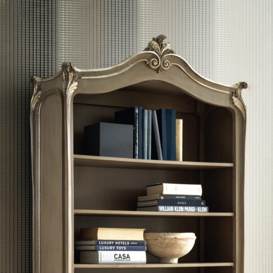 Rococo Inspired Bookcase The Rococo Inspired Bookcase, adorned with intricate carvings on top, is filled with a blend of stacked books and decorative items. A textured wall behind it further accentuates its vintage charm.