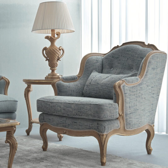 A vintage-style living room features a plush Rococo Style Armchair in blue, adorned with intricate carved wooden accents. Beside it, a coordinating wooden side table supports an ornate lamp with a pleated shade, all contrasted against a light grey backdrop.