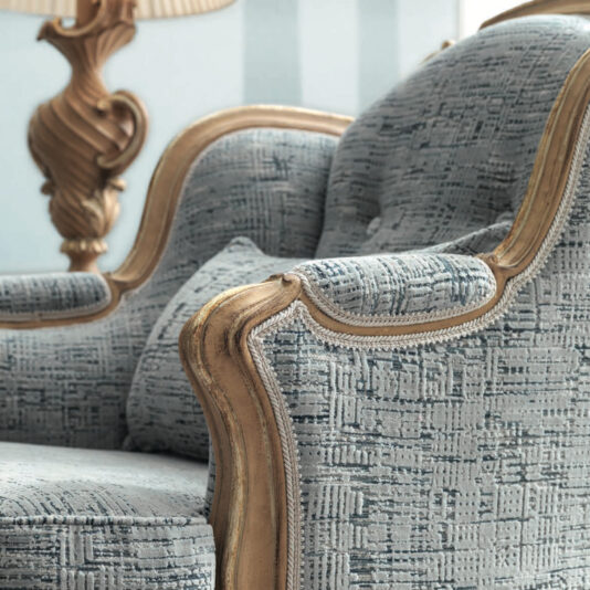 Rococo Style Armchair Close-up of the Rococo Style Armchair, showcasing its elegant vintage design with blue-patterned upholstery and a lavishly carved wooden frame. A coordinating decorative pillow is positioned on the seat, with a gracefully curved gold lamp providing ambient light in the background.