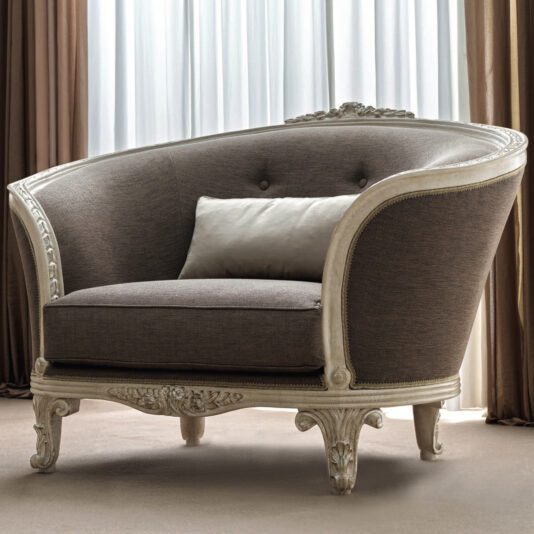 The Rococo Style Curved Armchair, featuring ornate wooden detailing and dark upholstery, sits gracefully in front of a window adorned with sheer curtains. Its elegant design cradles a light-colored, rectangular pillow that perfectly complements the neutral-toned drapes and flooring of the room.