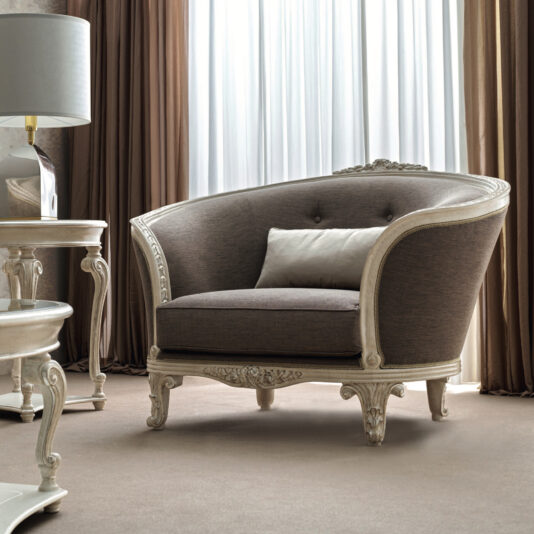 The Rococo Style Curved Armchair, featuring an ornate wooden frame and brown upholstery, sits gracefully in a softly lit room. Its curved features blend seamlessly with sheer and heavy curtains, while a small, ornate side table and lamp enhance the classic interior.