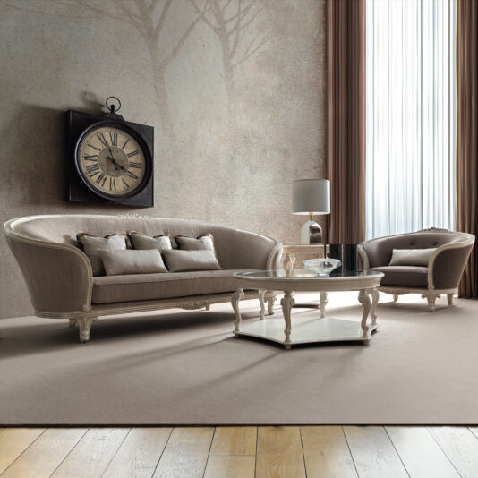 A stylish living room features a large beige sofa and the Rococo Style Curved Armchair. A glass coffee table sits at the center, while a lamp on a side table casts a warm glow. Above the couch hangs an elegant wall clock with Roman numerals, and sheer curtains drape over the tall window.