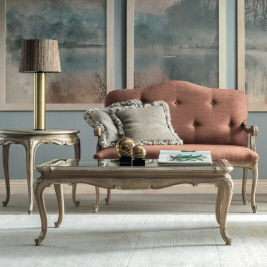 A vintage living room features a Small Rococo Style Day Sofa complemented by pink cushions and two grey pillows. A glass-top coffee table and a wooden side table with a lamp enhance its allure, while abstract paintings in muted tones on the walls contribute to an elegant and classic ambiance.