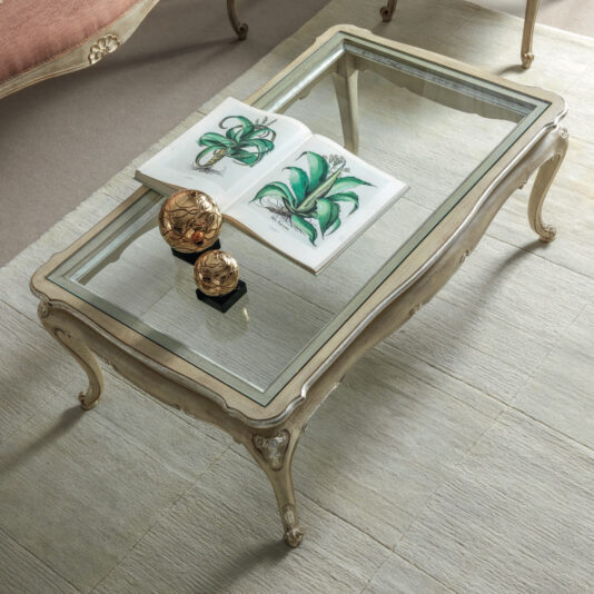 A glass-top coffee table displays a decorative open book filled with botanical illustrations of green plants. Two small, round golden ornaments are placed next to the book on top of a plush rug. Nearby, the setting exudes softness and elegance with a Small Rococo Style Day Sofa (Copy).