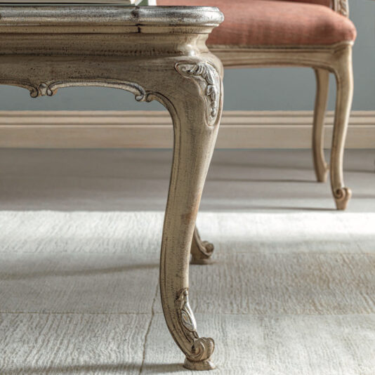 A close-up view of an intricately carved Rococo-style wooden table leg reveals its antique charm. In the background, a chair with a matching design and upholstered seat adds to the scene's elegance, set against a light-colored floor that highlights the furniture's timeless beauty, reminiscent of the Rococo Style Rectangular Glass Coffee Table.