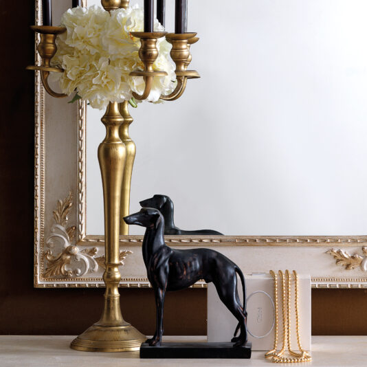 In a decorative setting, the Rococo Style Rectangular Wall Mirror, featuring lavish ornate detailing, is beautifully complemented by a brass candle holder with white flowers. Nearby, a black dog figurine, a small white book, and a string of pearls rest on the light-colored surface.