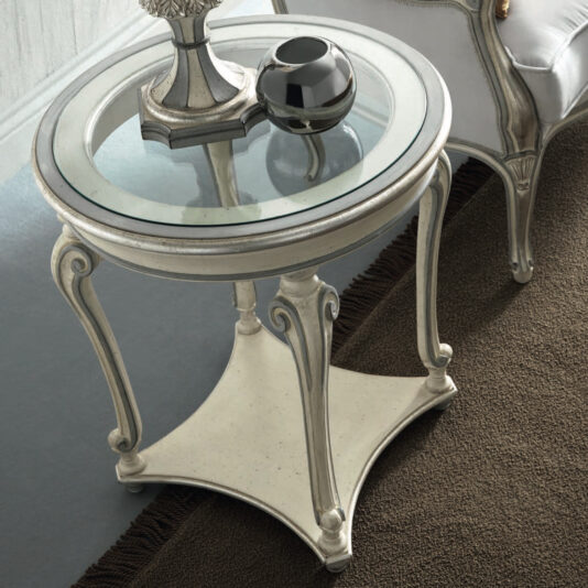 A Rococo Style Round Glass Side Table with an ornate metal base is situated on a brown carpet. The table is adorned with a silver decorative object and a black vase, while an upholstered chair can be seen nearby.
