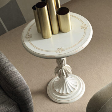 Rococo Style Round Side Table A Rococo Style Round Glass Side Table, featuring an ornate design and decorative pedestal base, is adorned with several tall, cylindrical brass vases. This elegant table rests on a beige carpet near a grey upholstered chair.