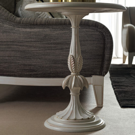 A Rococo Style Round Glass Side Table, featuring an ornate base with leaf motifs and a smooth round glass top, stands on a carpeted floor beside a gray sofa as sunlight streams in from a nearby window.