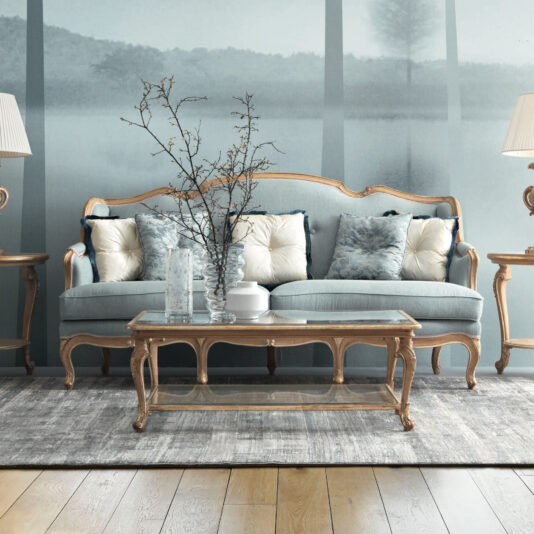 Rococo Style Sofa A chic living room showcases the Rococo Style Sofa, enhanced with light blue and white cushions, elegantly accompanied by a matching glass-top coffee table. A vase filled with branches adorns the table. The scene is completed by a plush rug and ambient floor lamps, crafting an inviting atmosphere.