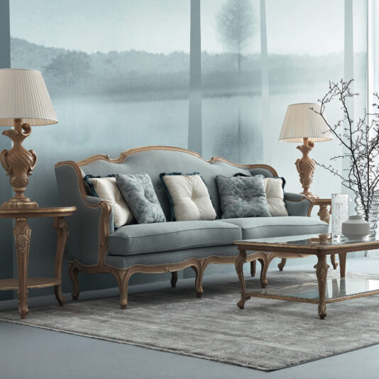 Rococo Style Sofa An elegant living room showcases a pastel blue Rococo Style Sofa, embellished with both patterned and plain pillows. The sofa is flanked by two ornate lamps on wooden side tables. In front of this setup, a glass coffee table rests on a gray rug, while misty landscape wallpaper adorns the background wall.